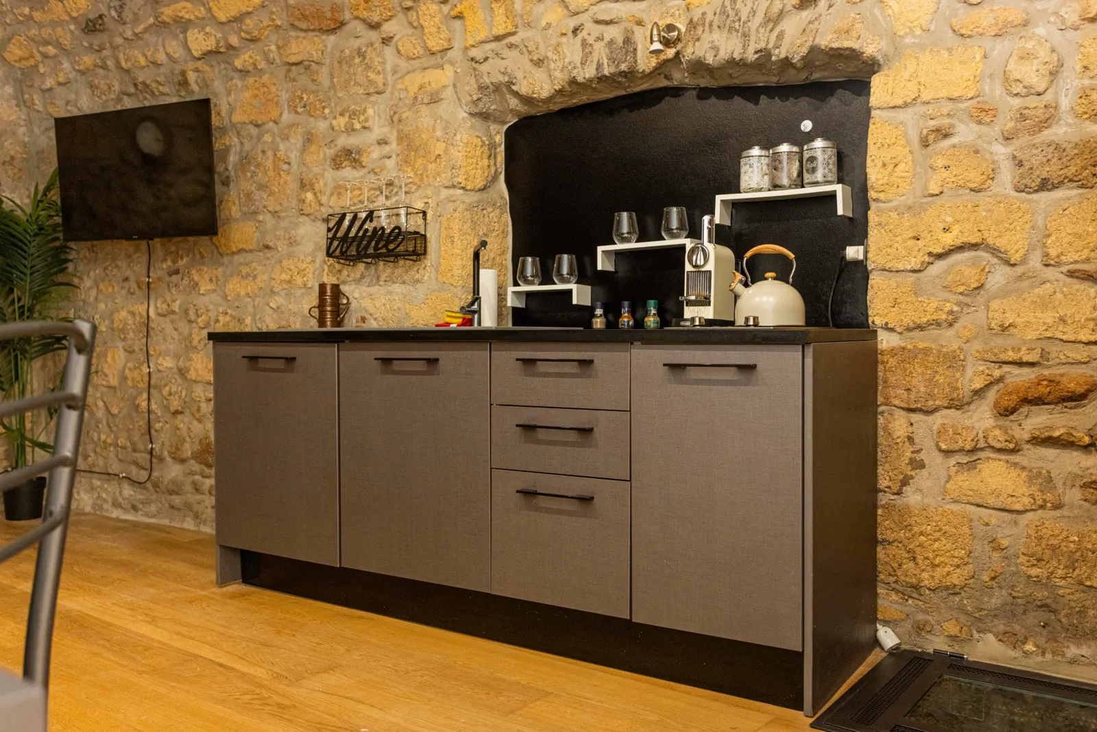 Angolo cucina del loft con mobili grigi, TV a parete e mensola Wine, nella parete di pietra