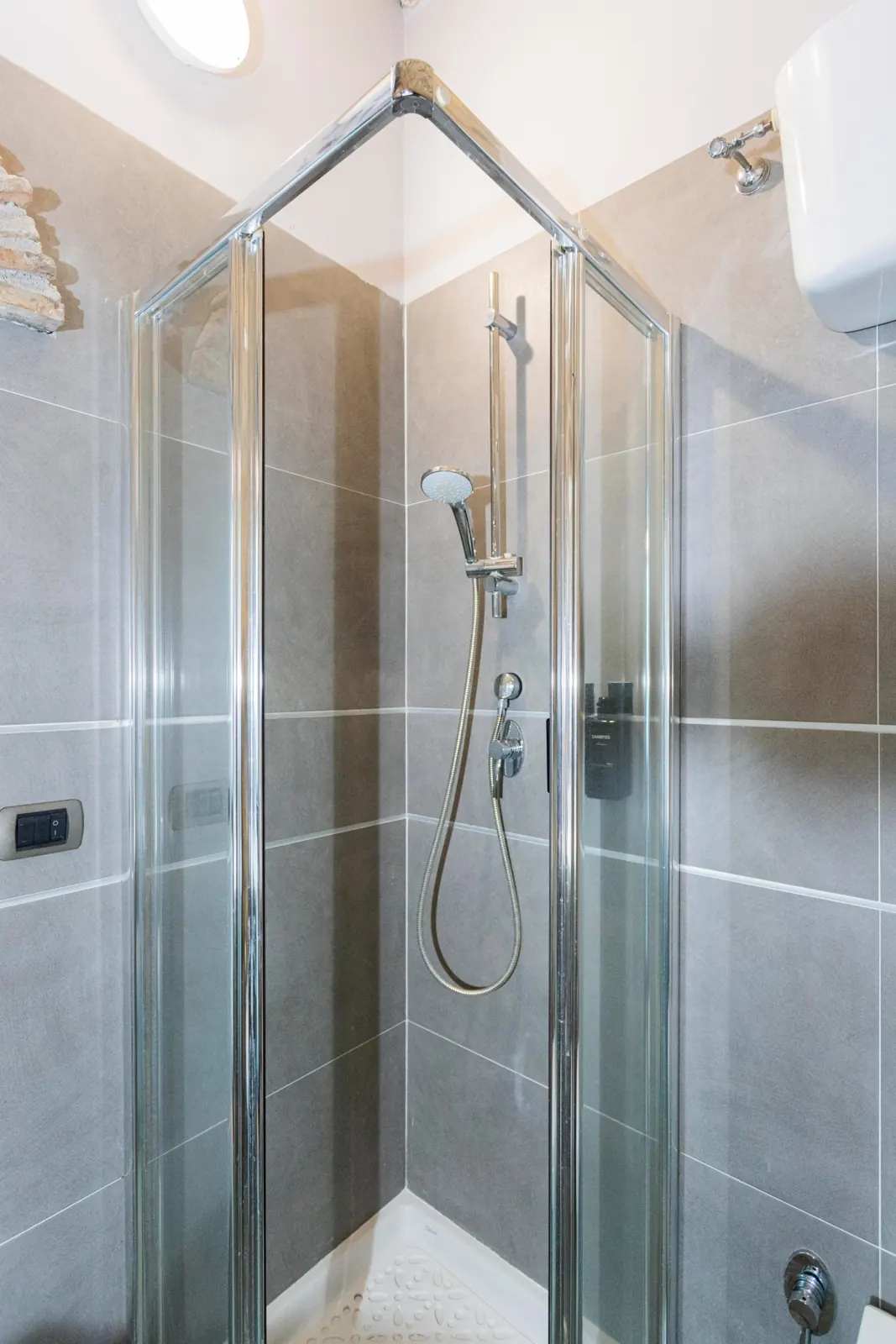 Doccia ad angolo con box in vetro, piastrelle grigie, doccino e dispenser di shampoo