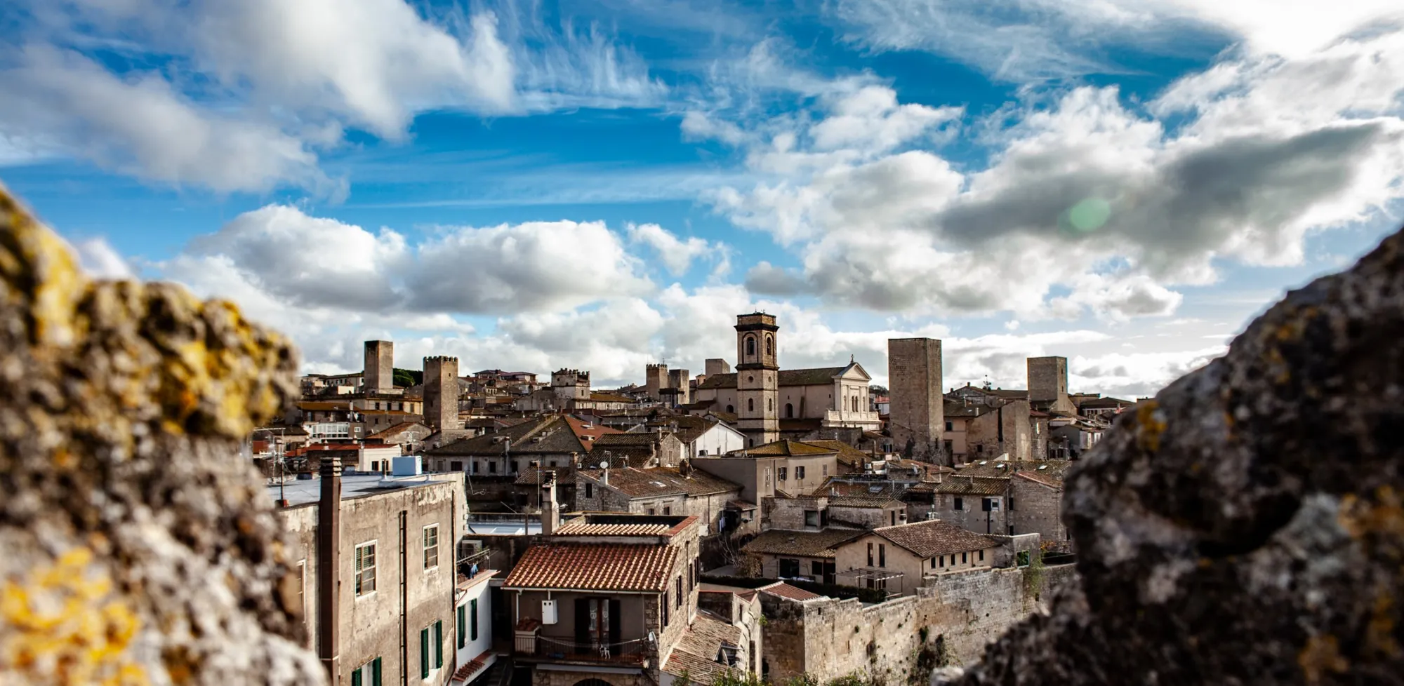 Tetti in coppo, torri medievali e campanile del centro storico di Tarquinia, visti tra due pietre sotto un cielo di nuvole